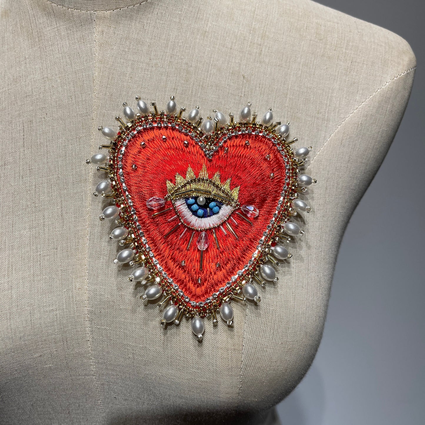 Front view of the 10x9cm Baroque Pearl Border hand-beaded sacred heart patch displayed on a beige linen dress form mannequin, showcasing the luxury outer layer of luminous baroque pearls, gold thread embroidery, and a central blue eye detail.
