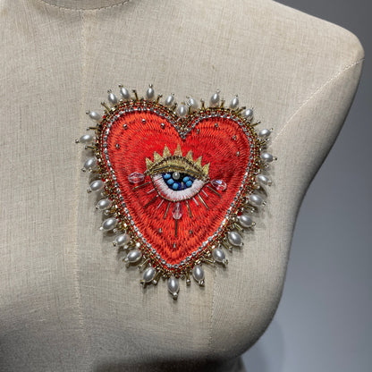 Front view of the 10x9cm Baroque Pearl Border hand-beaded sacred heart patch displayed on a beige linen dress form mannequin, showcasing the luxury outer layer of luminous baroque pearls, gold thread embroidery, and a central blue eye detail.