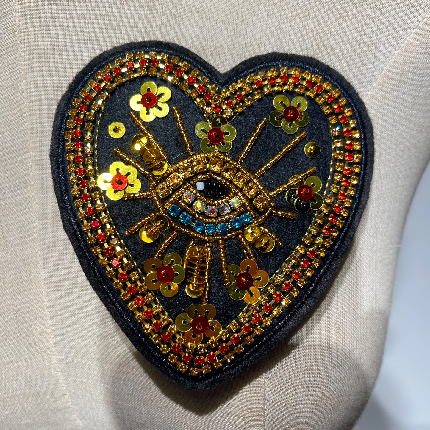 ⁠Close-up of a handmade black heart beaded embroidered patch featuring an evil eye motif with blue rhinestones, gold sequin flowers, and crystal borders.⁠