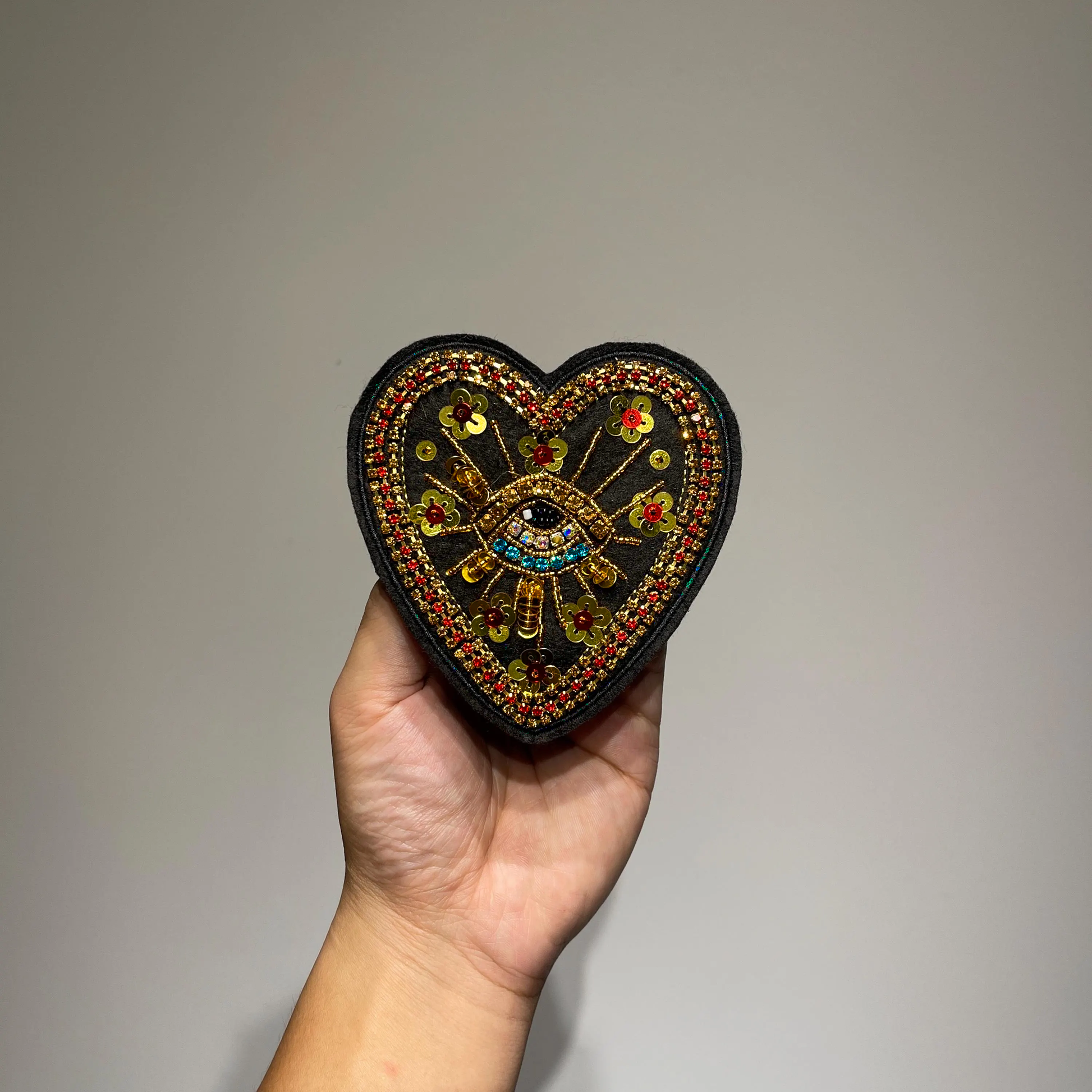 ⁠Hand holding a black heart-shaped evil eye beaded embroidered patch to demonstrate size and scale against a neutral background.⁠