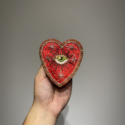 Close-up of a person holding the 10x9cm Celestial Starburst hand-beaded sacred heart patch, showcasing intricate red embroidery, metallic gold thread stars, and a central divine eye with green and glass micro-beads.