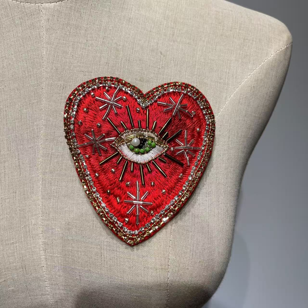 Front view of the 10x9cm Celestial Starburst hand-beaded sacred heart patch displayed on a beige linen dress form mannequin, highlighting the scattered gold metallic star burst rays and a central green beaded divine eye.