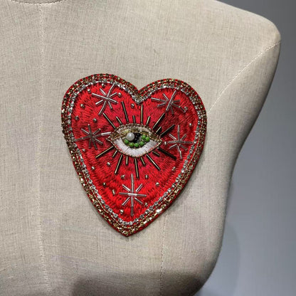 Front view of the 10x9cm Celestial Starburst hand-beaded sacred heart patch displayed on a beige linen dress form mannequin, highlighting the scattered gold metallic star burst rays and a central green beaded divine eye.