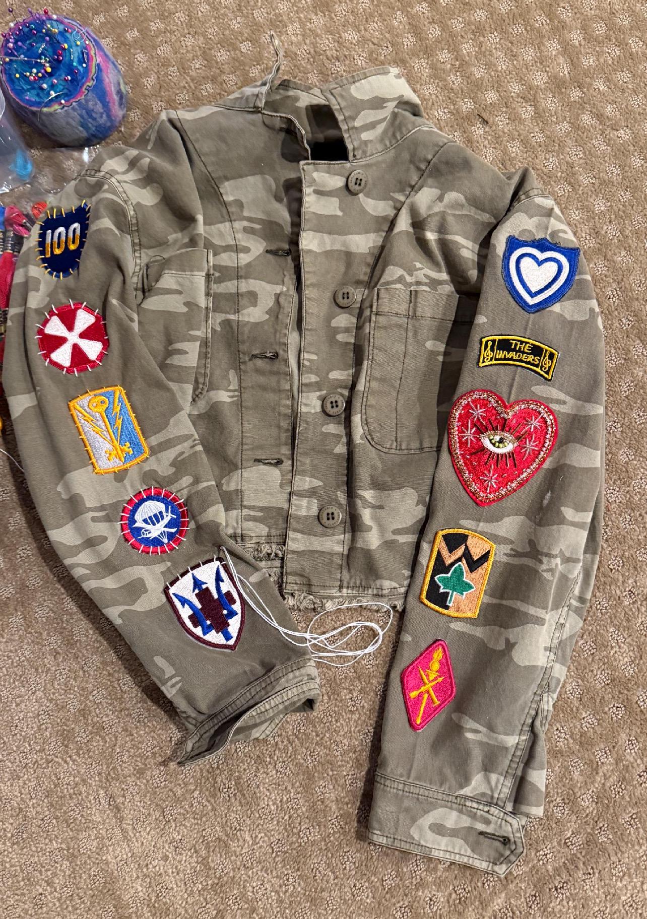 Flatlay view of a custom DIY camo utility jacket rework project, featuring various embroidered military and vintage style patches. Highlighted on the right sleeve is the premium 10x9cm Celestial Starburst hand-beaded sacred heart patch with intense red embroidery and gold thread details.
