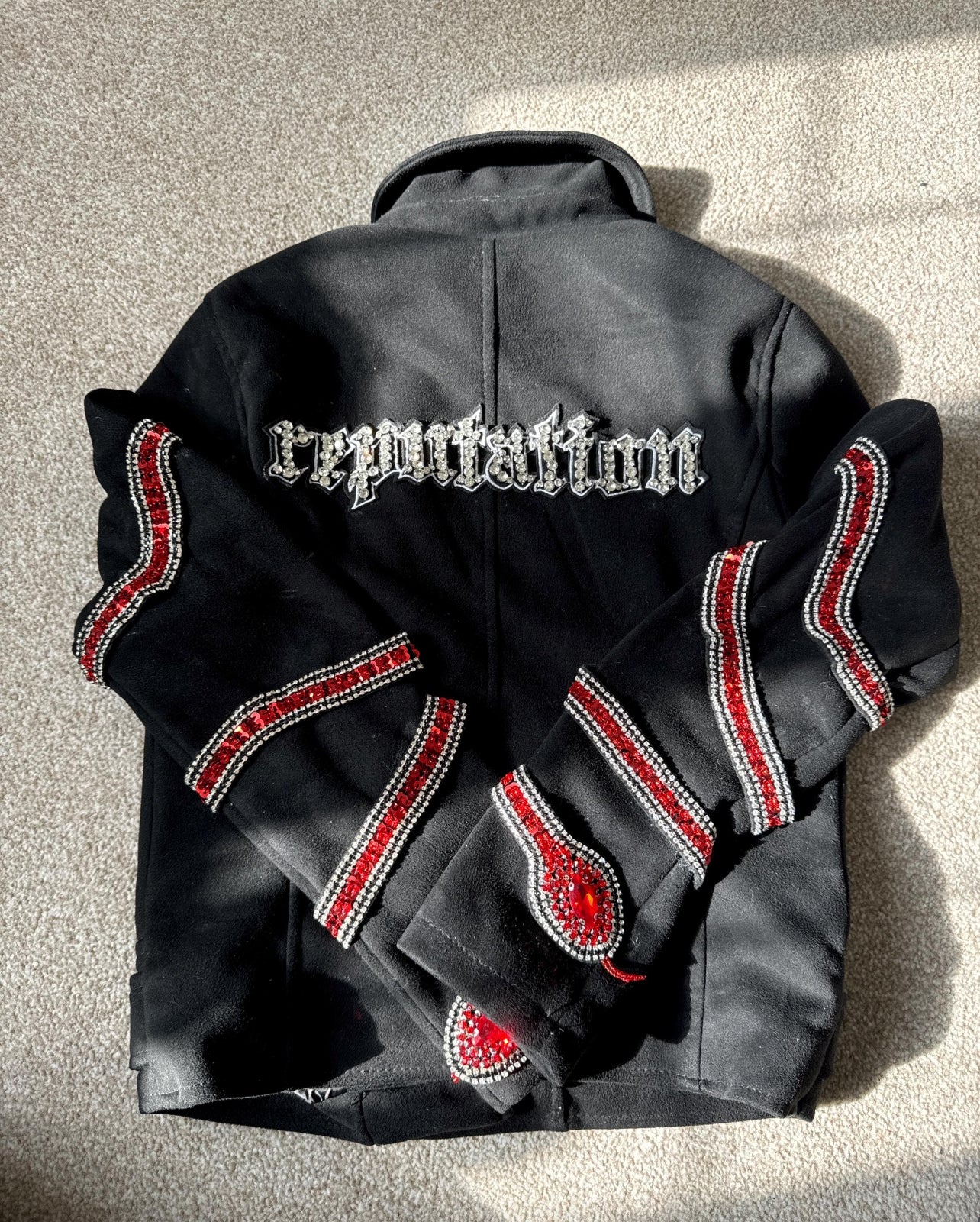 The back of a black jacket featuring "reputation" in silver gothic font embroidery across the shoulders, with red and silver rhinestone snake patterns wrapping around the sleeves.⁠