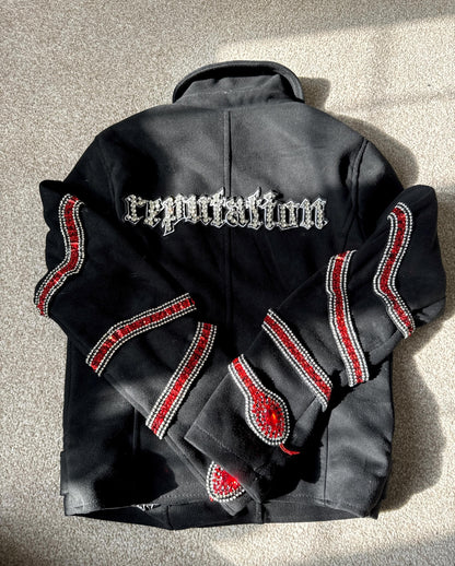 The back of a black jacket featuring "reputation" in silver gothic font embroidery across the shoulders, with red and silver rhinestone snake patterns wrapping around the sleeves.⁠