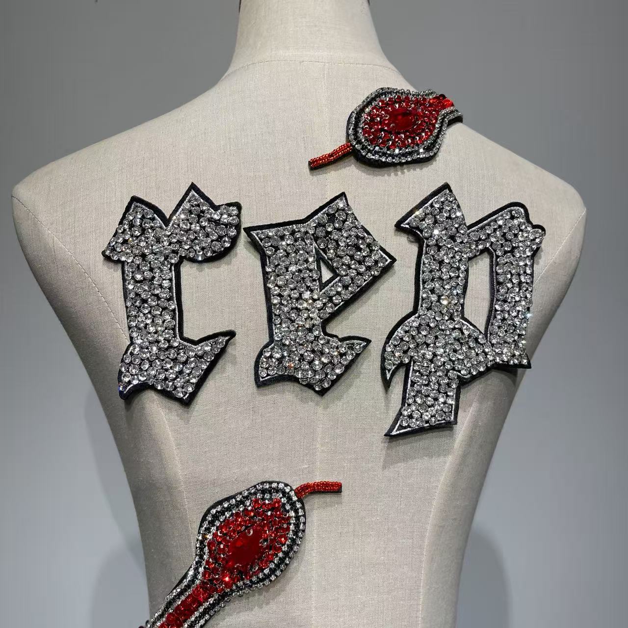 A close-up of a cream-colored mannequin torso featuring glittering rhinestone patches. The center displays large Gothic-style "REP" letters encrusted with silver crystals and black borders. Two red and black rhinestone snake head patches are positioned above and below the text