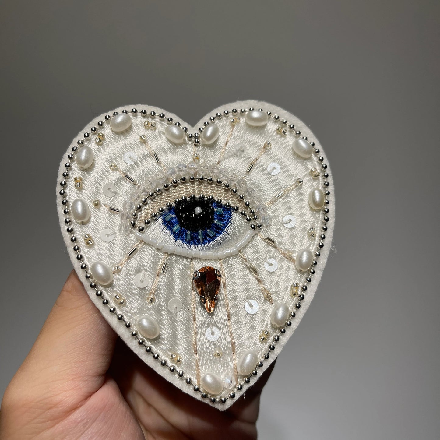 Hand holding a handmade white evil eye beaded heart patch with embroidery, pearls, sequins, and crystal pendant - 9.5x8cm.