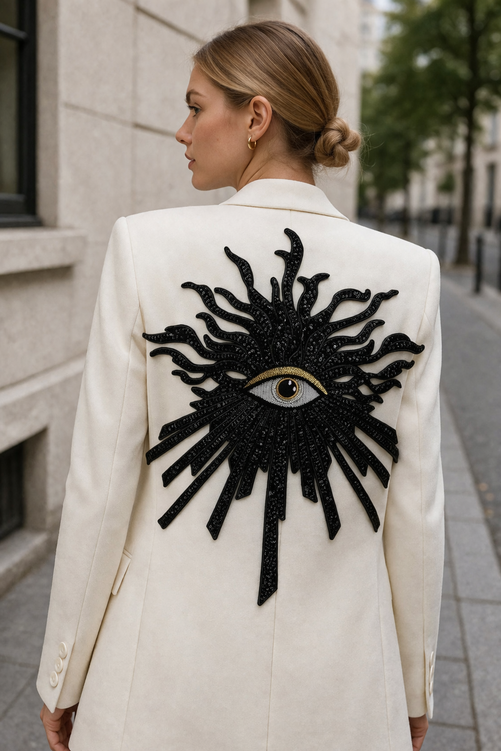 Chic blonde model wearing a white premium blazer jacket featured with an oversized black evil eye sequin back patch on a street, luxury custom streetwear styling ideas.