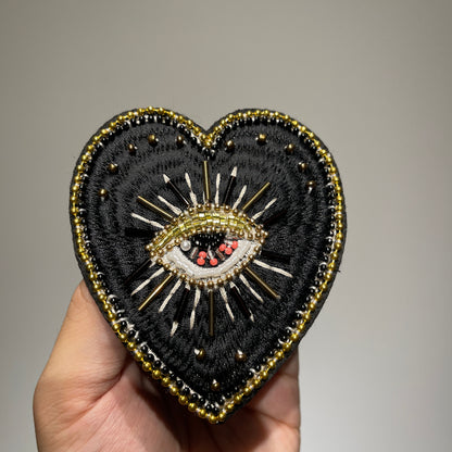 ⁠Close-up view of artisanal hand-stitched black and gold embroidery with pearl and beadwork on an evil eye heart patch - 9x8.5cm.⁠