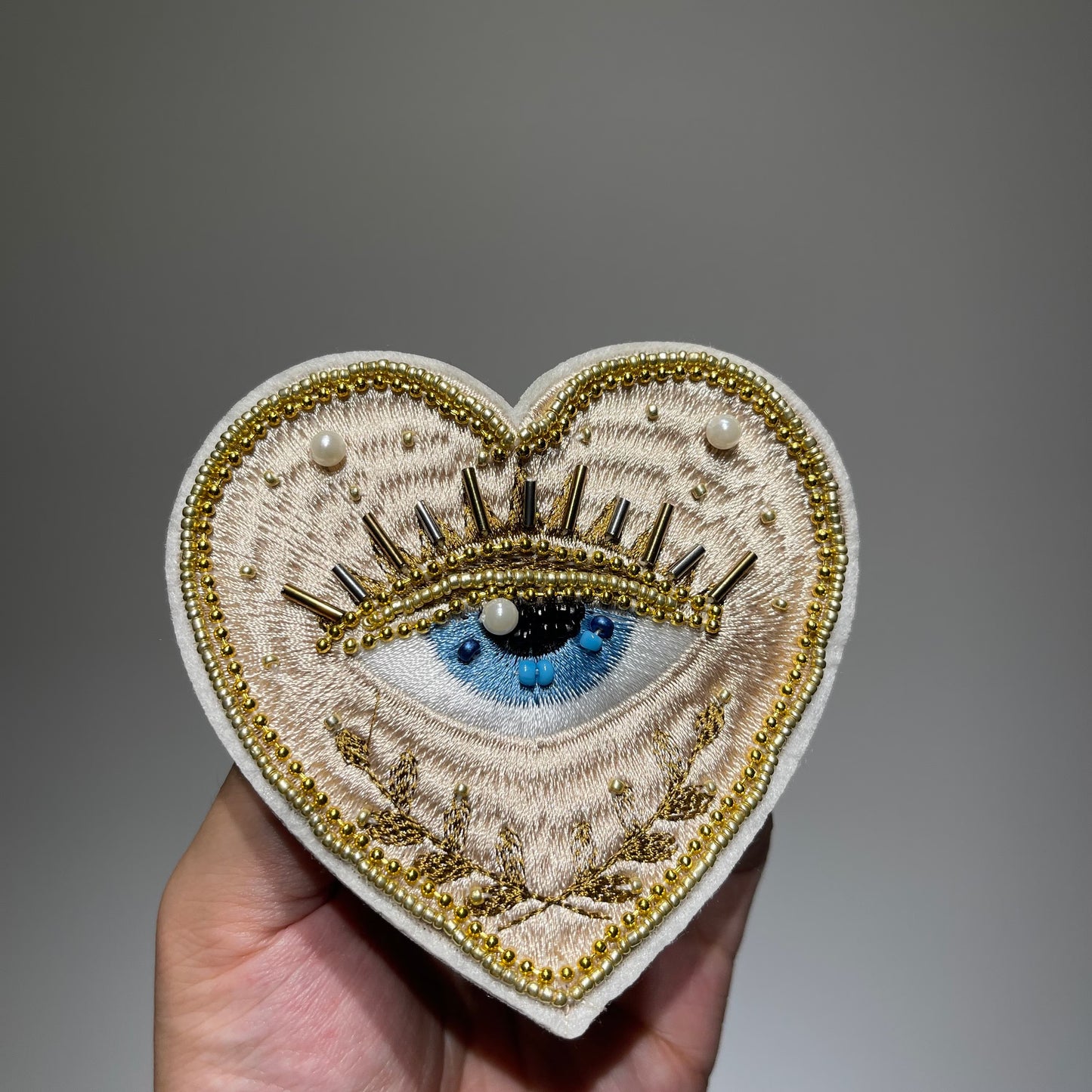 ⁠Close-up view of artisanal hand-stitched gold embroidery and pearl beadwork on a 10cm evil eye heart patch applique.⁠