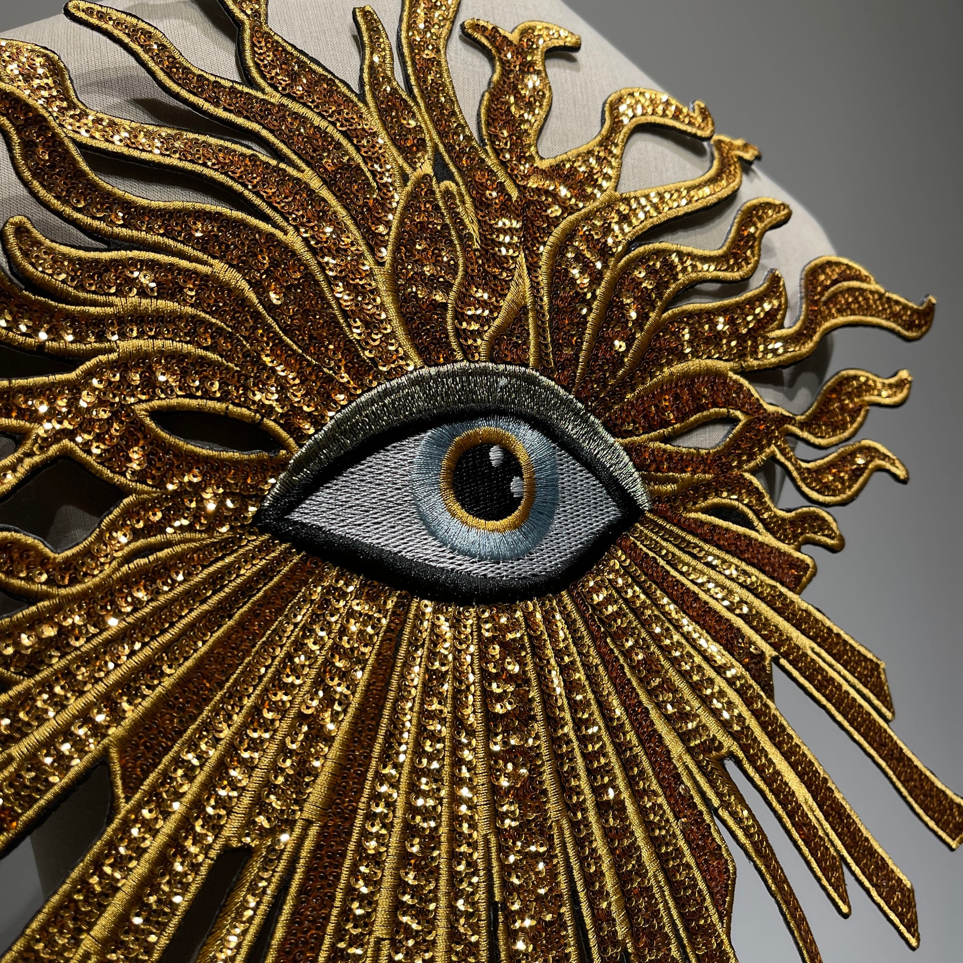 Side angle close-up of premium gold evil eye applique patch, showing dimensional 3D embroidery texture and reflective light on metallic sequins