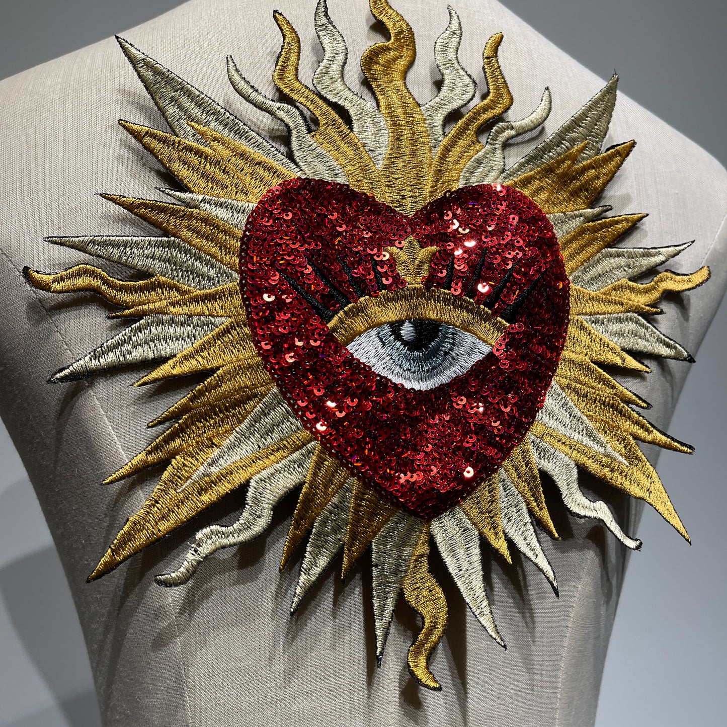 Close-up detail of luxury red sacred heart patch, showcasing shimmering ruby sequins, metallic gold and silver sunburst embroidery threads, and exquisite artisan needlework.