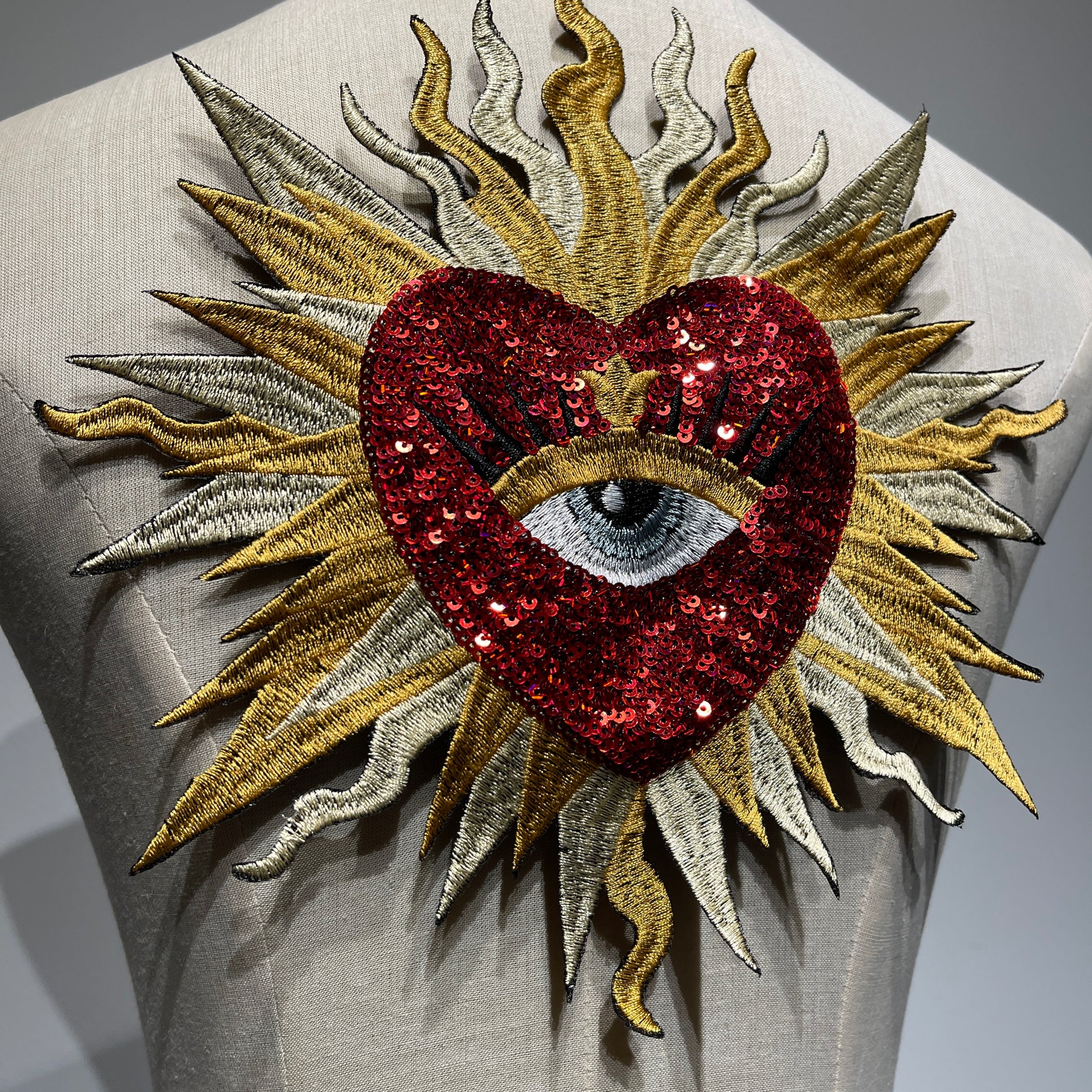 Close-up detail of luxury red sacred heart patch, showcasing shimmering ruby sequins, metallic gold and silver sunburst embroidery threads, and exquisite artisan needlework.
