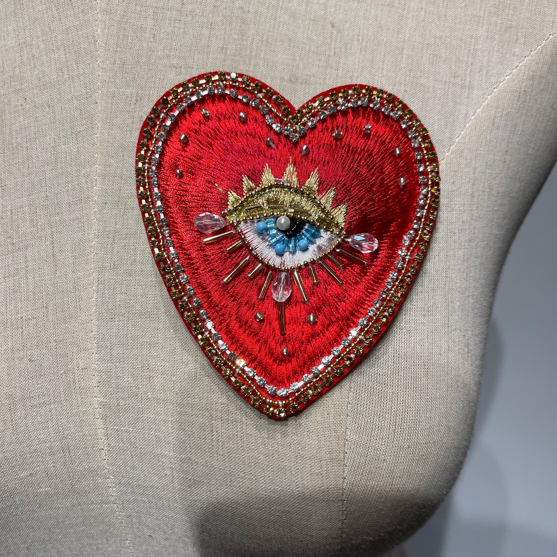 Close-up detail of the Radiant Gold Sunburst hand-beaded sacred heart patch (10x9cm), featuring intricate red embroidery, a mystical divine eye centerpiece, and shimmering gold metallic sunburst rays, displayed on a textured linen background.