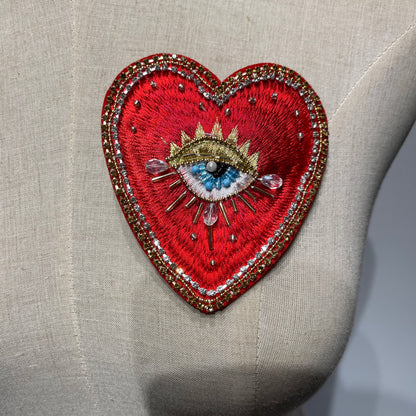 Close-up detail of the Radiant Gold Sunburst hand-beaded sacred heart patch (10x9cm), featuring intricate red embroidery, a mystical divine eye centerpiece, and shimmering gold metallic sunburst rays, displayed on a textured linen background.