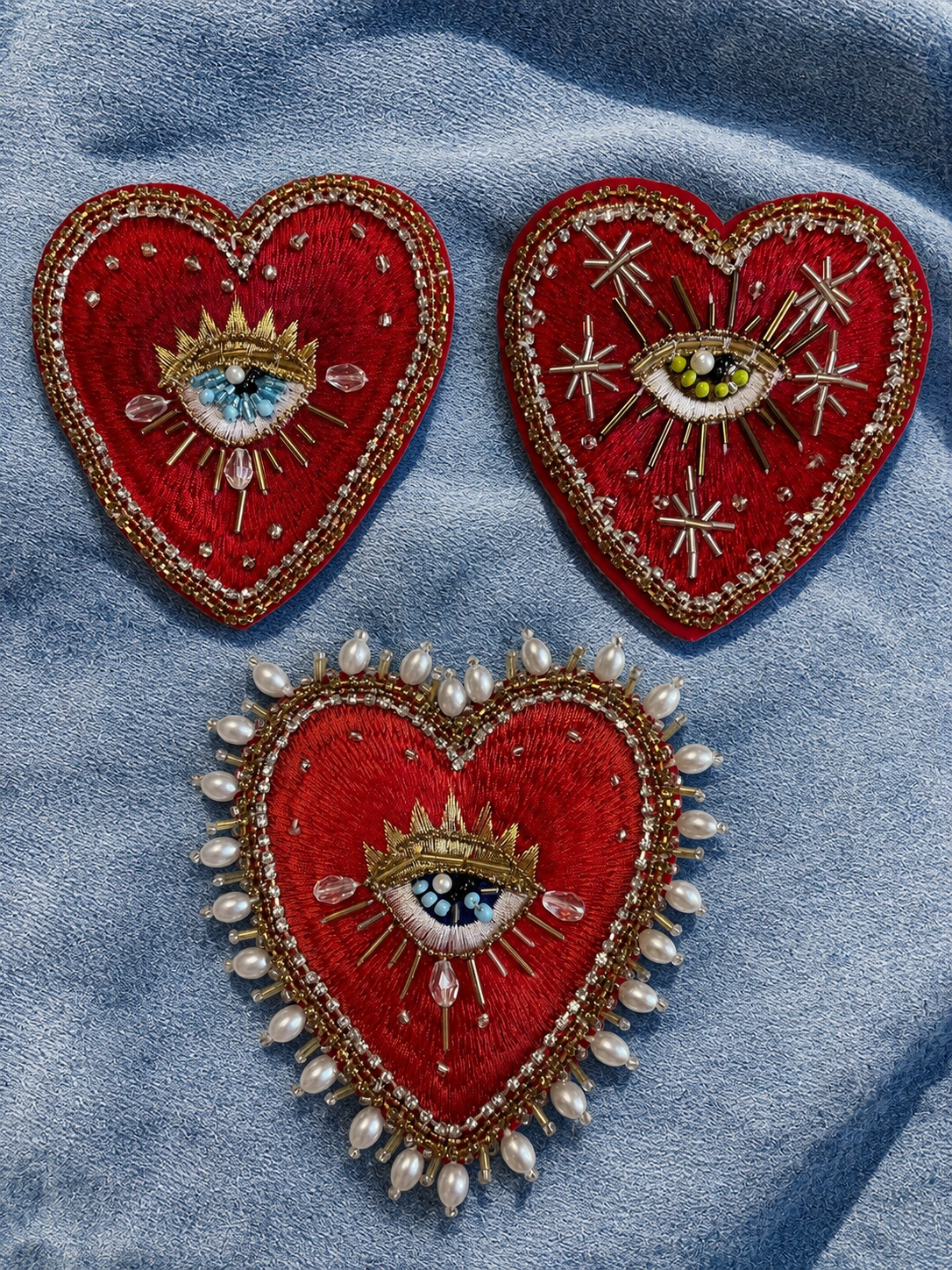 Flatlay view of three distinct 10x9cm hand-beaded sacred heart patches—Sunburst, Celestial, and Baroque Pearl Border styles—displayed together on a blue denim background, showcasing intricate red embroidery, gold threads, and divine eye details for custom apparel rework.
