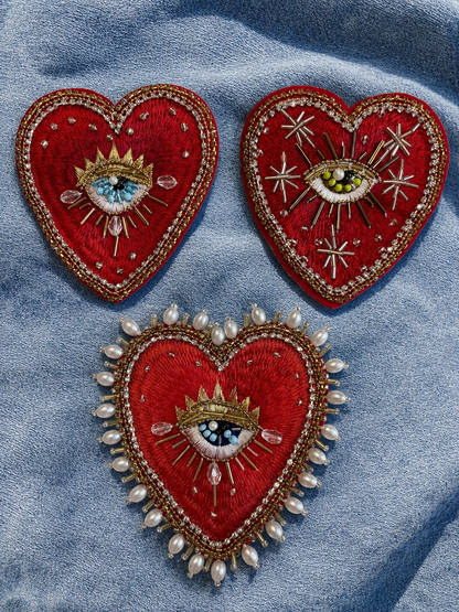 Flatlay view of three distinct 10x9cm hand-beaded sacred heart patches—Sunburst, Celestial, and Baroque Pearl Border styles—displayed together on a blue denim background, showcasing intricate red embroidery, gold threads, and divine eye details for custom apparel rework.